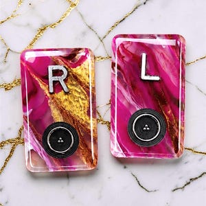 May include: Two rectangular resin pieces with a vibrant pink and gold abstract design. One features a silver "R" and the other a silver "L", both with a black button detail. The background is a marble surface.