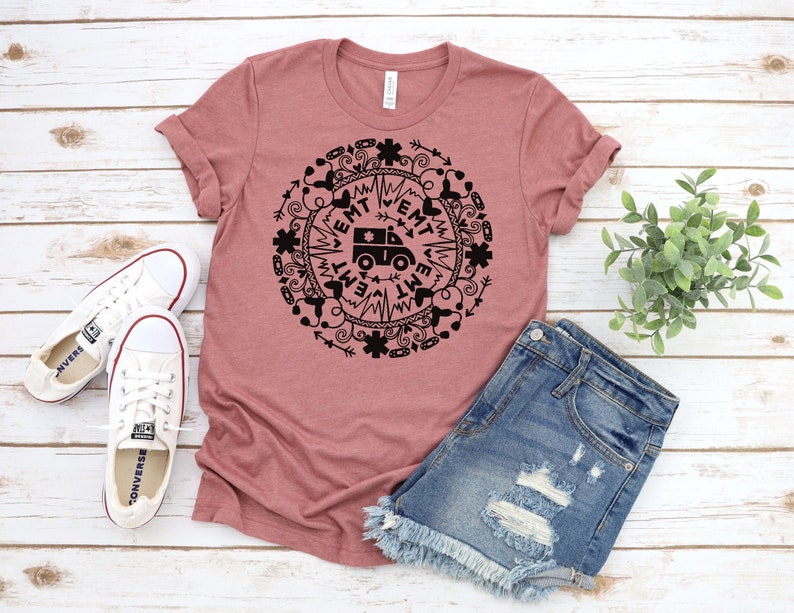 May include: A mauve t-shirt with a black graphic design of an ambulance surrounded by a circle of medical symbols and the word "EMT" repeated around the circle.