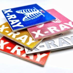May include: Five different colored rectangular signs with the text "X-RAY" printed in white on each. The signs are stacked on top of each other, with the blue sign on top and the red sign on the bottom. The signs are made of a textured material.