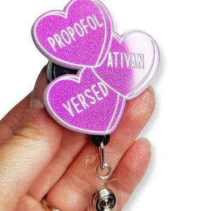 May include: A glittery purple badge reel with three heart-shaped charms. Each charm has a white outline and text: "PROPOFOL", "ATIVAN", and "VERSED". The reel has a black backing, a clear plastic strap, and a metal clip.