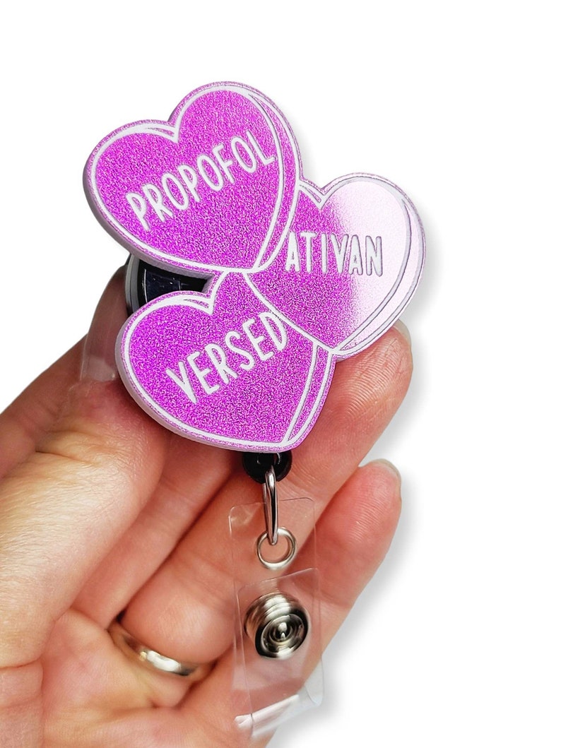 May include: A glittery purple badge reel with three heart-shaped charms. Each charm has a white outline and text: "PROPOFOL", "ATIVAN", and "VERSED". The reel has a black backing, a clear plastic strap, and a metal clip.