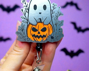 May include: A Halloween-themed badge reel featuring a silver ghost character hugging an orange jack-o'-lantern. The design includes floral accents and is attached to a black retractable reel with a clear plastic clip. The background is purple with bat silhouettes.