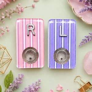 May include: Two rectangular acrylic blocks featuring vertical stripes in pink and purple. Each block has a silver-toned letter, R on the pink block and L on the purple block, positioned above a circular metallic indentation with a small black dot.