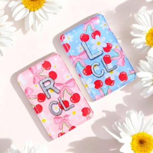 May include: Two small rectangular tiles with a cherry and bow pattern. The tiles are pink and blue and have the letters "RCL" and "LCL" on them.