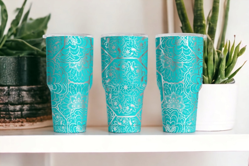 May include: Three turquoise tumblers with silver mandala designs. The tumblers are on a white shelf.