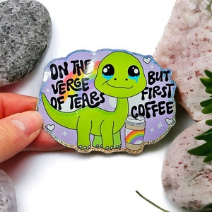 K&ouml;nnte beinhalten: Ein holografischer Aufkleber mit einem weinenden gr&uuml;nen Dinosaurier, der eine Kaffeetasse mit einem Regenbogen h&auml;lt. Der Text lautet "On the verge of tears but first coffee".