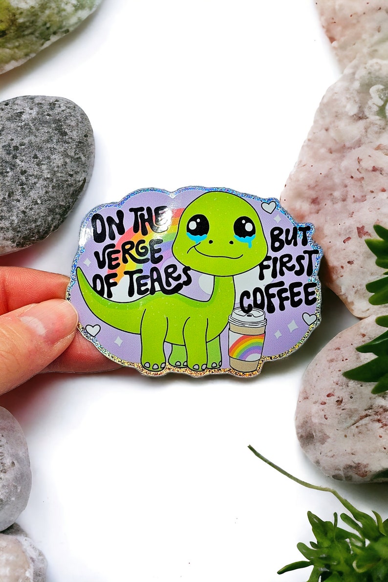 K&ouml;nnte beinhalten: Ein holografischer Aufkleber mit einem weinenden gr&uuml;nen Dinosaurier, der eine Kaffeetasse mit einem Regenbogen h&auml;lt. Der Text lautet "On the verge of tears but first coffee".