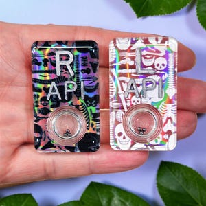 May include: Two rectangular, decorative buttons with the letters "RAPI" on them. One button is black with a holographic design and the other is white with a skeleton design. Each button has a circular, clear center.