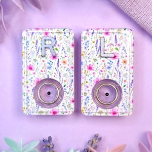 May include: Two rectangular resin hearing aid covers featuring a colorful wildflower pattern with pink, blue, and yellow blossoms. Each cover displays a silver letter, R on the left and L on the right, with a circular purple center component.
