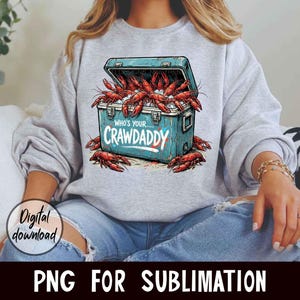 Puede incluir: Una sudadera gris claro con un gr&aacute;fico de una nevera azul desbordante de cangrejos rojos. El texto "WHO'S YOUR...CRAWADDY" est&aacute; impreso en la nevera. Tambi&eacute;n se ven las palabras "Digital download" y "PNG FOR SUBLIMATION".