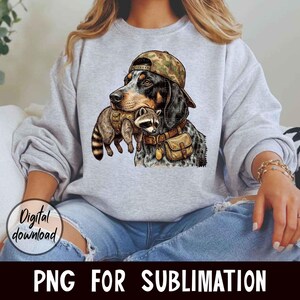 May include: A gray sweatshirt featuring a graphic of a dog wearing a camouflage cap and holding a raccoon. The dog has a brown leather collar and a small bag. The words "Digital download" and "PNG FOR SUBLIMATION" are also visible.
