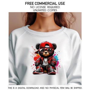 Pu&ograve; includere: Una felpa bianca con una grafica colorata di un orso dei cartoni animati che indossa un cappello rosso, occhiali da sole e una catena d'oro. L'orso &egrave; seduto su uno sfondo di schizzi colorati.