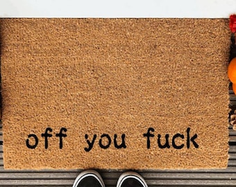 May include: Brown coir doormat with the words "off you fuck" in black. The mat is surrounded by pumpkins, pinecones, and fall foliage. Black and white sneakers are at the bottom.
