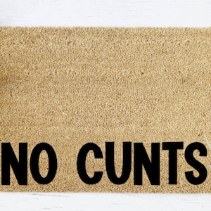 May include: A rectangular coir doormat in a tan colour. The words "NO CUNTS" are printed in bold black letters across the center. The mat is surrounded by white wood and decorative greenery.