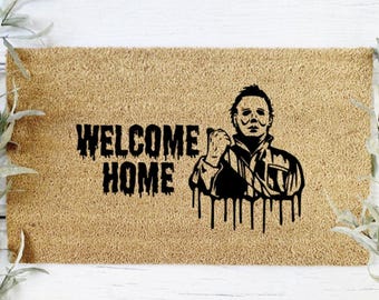 May include: A rectangular coir doormat with the words "WELCOME HOME" in dripping black letters. A black silhouette of a figure with a mask and knife is on the right. The mat is a light brown color.