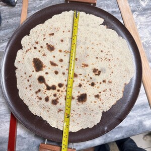 May include: A large, round, light-colored flatbread with brown spots, resting on a dark brown griddle. A yellow measuring tape shows the bread is approximately 16 inches in diameter. Wooden rolling pins and a red-handled tool are nearby.