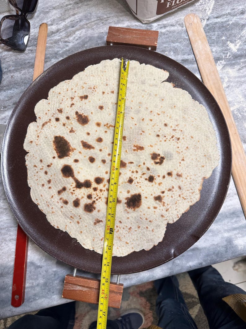May include: A large, round, light-colored flatbread with brown spots, resting on a dark brown griddle. A yellow measuring tape shows the bread is approximately 16 inches in diameter. Wooden rolling pins and a red-handled tool are nearby.