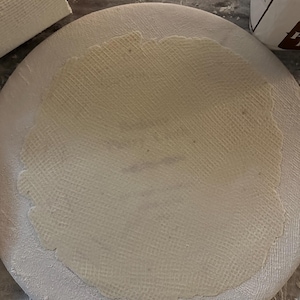 May include: A round, off-white, textured dough is centered on a white, circular surface dusted with flour. A red-handled rolling pin rests nearby. The scene suggests baking or food preparation.