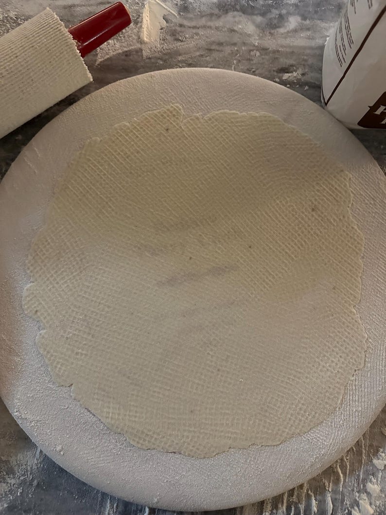 May include: A round, off-white, textured dough is centered on a white, circular surface dusted with flour. A red-handled rolling pin rests nearby. The scene suggests baking or food preparation.