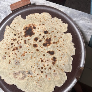 May include: A round, light-colored flatbread with dark brown spots, resting on a dark brown plate. The edges of the flatbread are slightly uneven. The plate is on a light-colored surface.