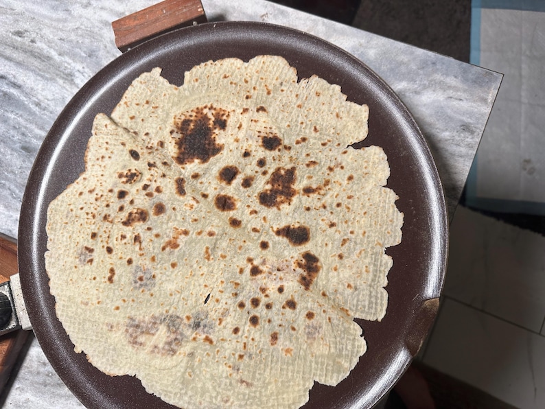 May include: A round, light-colored flatbread with dark brown spots, resting on a dark brown plate. The edges of the flatbread are slightly uneven. The plate is on a light-colored surface.