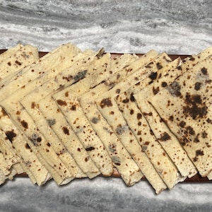 May include: A stack of thin, light-colored flatbreads with dark brown spots, arranged on a wooden cutting board. The flatbreads are cut into triangles and fanned out, showcasing their texture and color variations. The background is a marble-like surface.