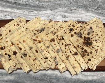 May include: A stack of thin, light-colored flatbreads with dark brown spots, arranged on a wooden cutting board. The flatbreads are cut into triangles and fanned out, showcasing their texture and color variations. The background is a marble-like surface.