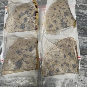 May include: Four individually packaged, triangular-shaped food items. The food has a light brown color with dark spots, and is sealed in clear plastic. The packaging is on a light gray surface.
