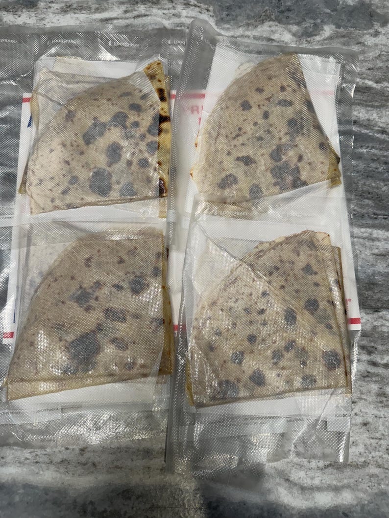 May include: Four individually packaged, triangular-shaped food items. The food has a light brown color with dark spots, and is sealed in clear plastic. The packaging is on a light gray surface.