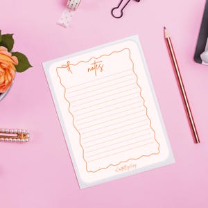 Può includere: Un blocco note con sfondo color crema e bordo dorato decorativo. La parola "notes" è scritta in oro nella parte superiore. Il blocco note ha pagine a righe e il testo "#CraftedByKing" in basso. Una matita dorata poggia sulla superficie rosa.