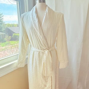 May include: A white, long-sleeved robe with a satin-like finish and a floral pattern. The robe has a shawl collar and a tie belt.