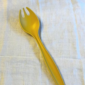 May include: A single, bright yellow serving fork with three tines. The fork is made of plastic and has a long, slender handle. The fork is lying on a white, textured tablecloth. The background includes a floral design.