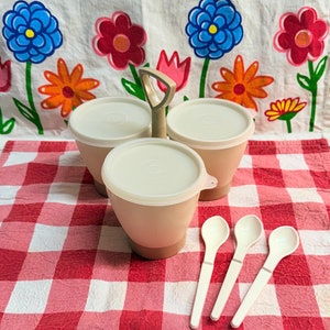 May include: A set of three beige plastic containers with lids and a beige plastic handle. The containers are on a red and white checkered tablecloth. There are three white plastic spoons next to the containers.