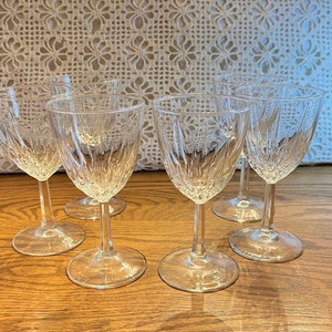 May include: Six clear glass wine glasses with a cut glass design. The glasses are arranged in a semi-circle on a wooden surface.