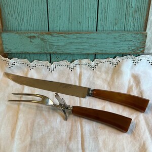 May include: A vintage carving knife and fork set with brown handles. The stainless steel blade of the knife has text. The fork has a metal guard. The set is on a white cloth with a decorative edge.