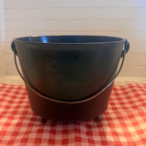 May include: A black cast iron Dutch oven with a metal handle. The pot has three small feet and sits on a red and white checkered tablecloth. The background is a white wall.