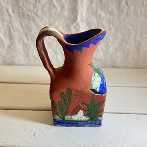 May include: A small terracotta pitcher with a blue, green, and white painted design. The pitcher features a cactus, a person wearing a hat, and a blue river.