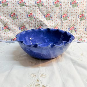 May include: A handmade, cobalt blue ceramic bowl with a ruffled edge. The bowl has a glossy finish and is set on a white surface with a floral patterned background. This decorative bowl is ideal for serving or display.