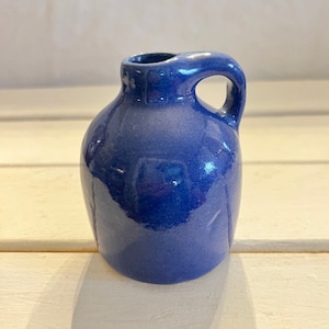 May include: A small, blue ceramic pitcher with a handle. The pitcher is glazed and has a shiny finish.