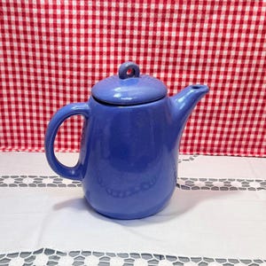May include: A blue ceramic teapot with a matching lid. The teapot has a curved handle and a spout. The lid has a small loop on top. The teapot is sitting on a white surface with a red and white checkered background.
