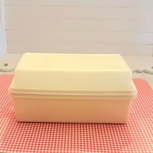 May include: A rectangular, two-tone plastic container with a lid. The base is a light peach color, and the lid is a creamy off-white. The container sits on a red and white checkered surface, suggesting a kitchen or food storage item.