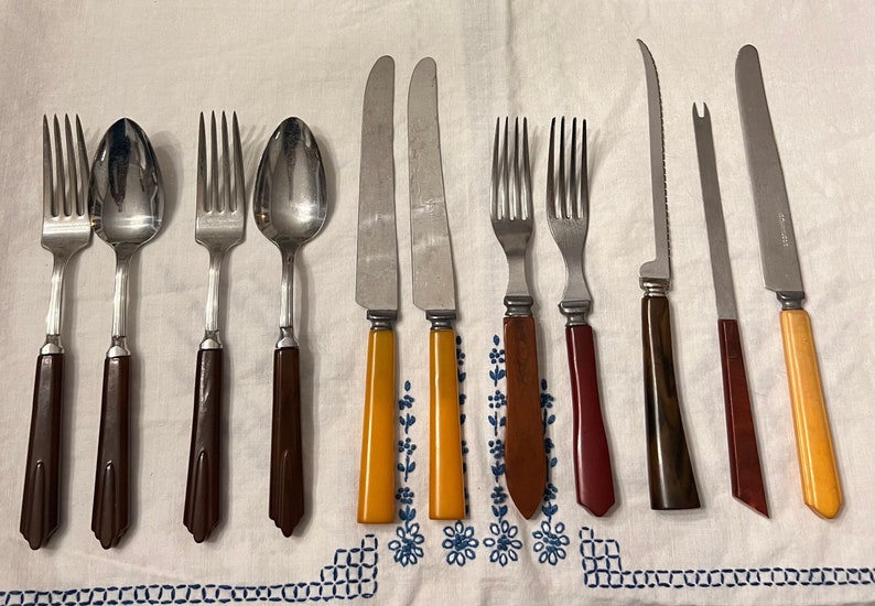 May include: A set of vintage cutlery with brown, red, and yellow plastic handles. The set includes forks, spoons, and knives.