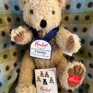 May include: A beige teddy bear wearing a blue ribbon around its neck. The bear has a tag that reads "Hamleys Est. London 1760 The Finest Toy Shop in the World Charles Heritage Bears Collection".