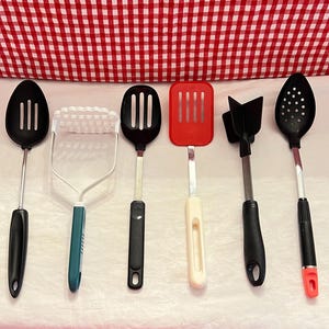 May include: A set of six kitchen utensils, including a potato masher, a slotted spoon, a spatula, and three solid spoons. The utensils are all black with black handles, except for the spatula, which is red with a white handle.