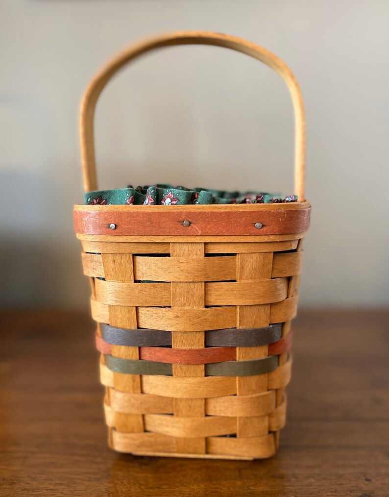 Pode incluir: Uma cesta tecida com uma al&ccedil;a de madeira e um forro de tecido verde floral. A cesta tem formato retangular com faixas cinza, laranja e verde tecidas na madeira natural. A borda &eacute; adornada com uma faixa marrom.