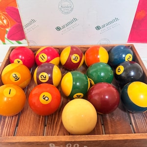 May include: A collection of billiard balls in a wooden tray. The balls are in various colours, including red, yellow, green, and blue, with white numbers. The tray is on a white surface.
