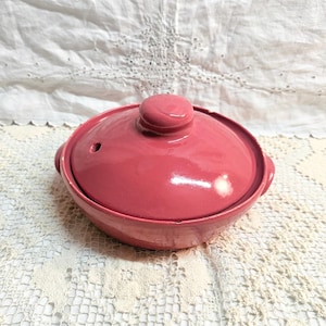 May include: A round, pink ceramic cooking pot with a matching lid. The lid has a small knob and a vent hole. The pot has two small handles on either side. The pot is sitting on a white lace tablecloth.