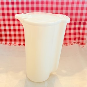 May include: A white plastic pitcher with a lid. The pitcher has a curved handle and a spout for easy pouring. The lid has a small tab for opening and closing. The pitcher is sitting on a white surface with a red and white checkered background.