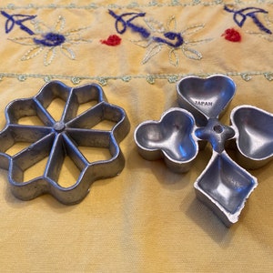 May include: Two vintage metal cookie cutters, one shaped like a flower with eight points and the other shaped like a four-leaf clover. The clover cutter has the word "JAPAN" stamped on it.