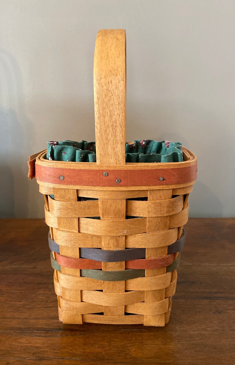Pode incluir: Cesto tecido retangular com al&ccedil;a de madeira e faixas decorativas em tons de castanho, laranja e verde. O interior &eacute; forrado com tecido floral verde. Um item de armazenamento decorativo.
